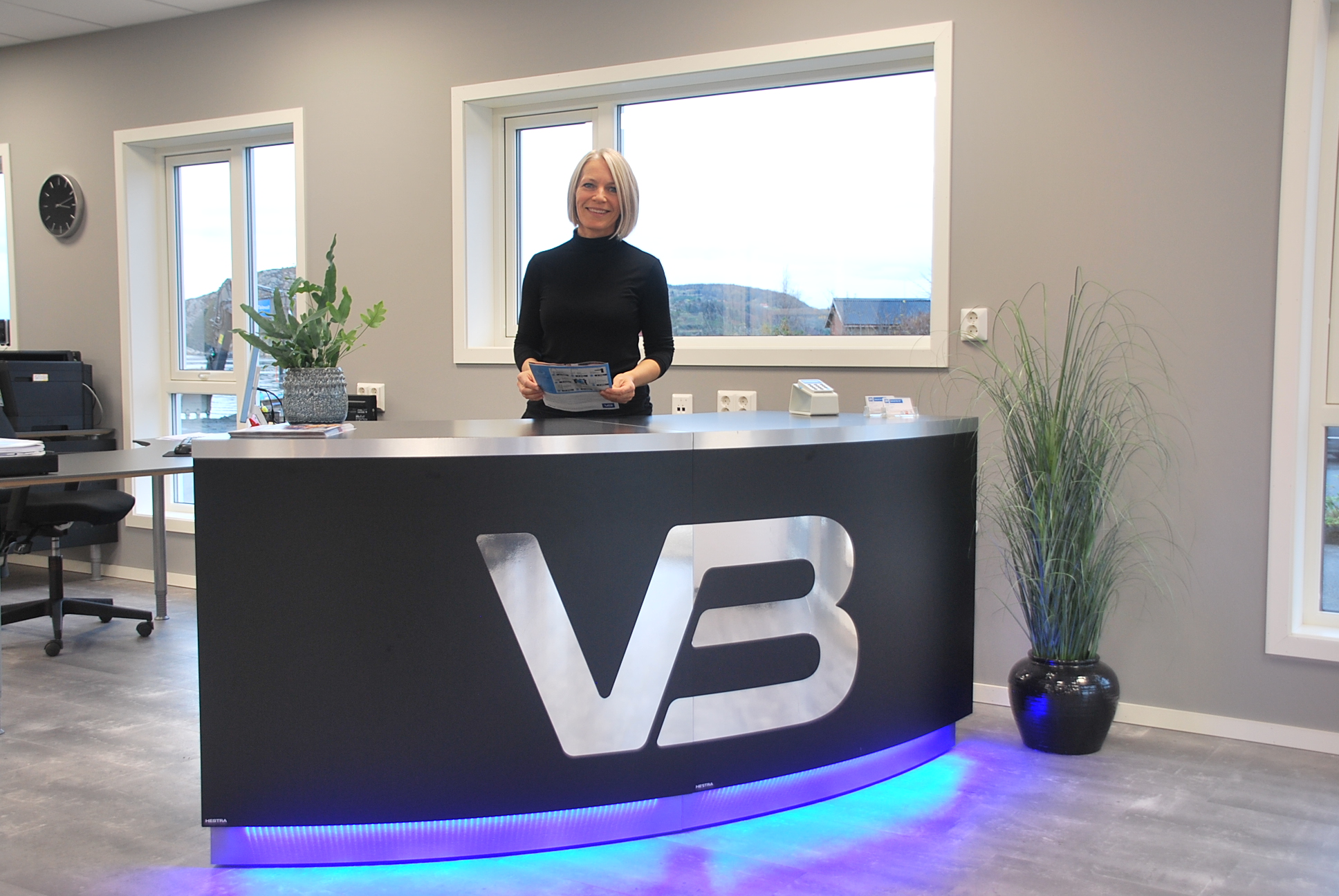 Resepsjonen hos VB-rørlegger Hatlevik Rør: Buet disk med brun front og VB-logo i sølv, kvinne bak disken og antydning til fjellutsikt utenfor vinduene.